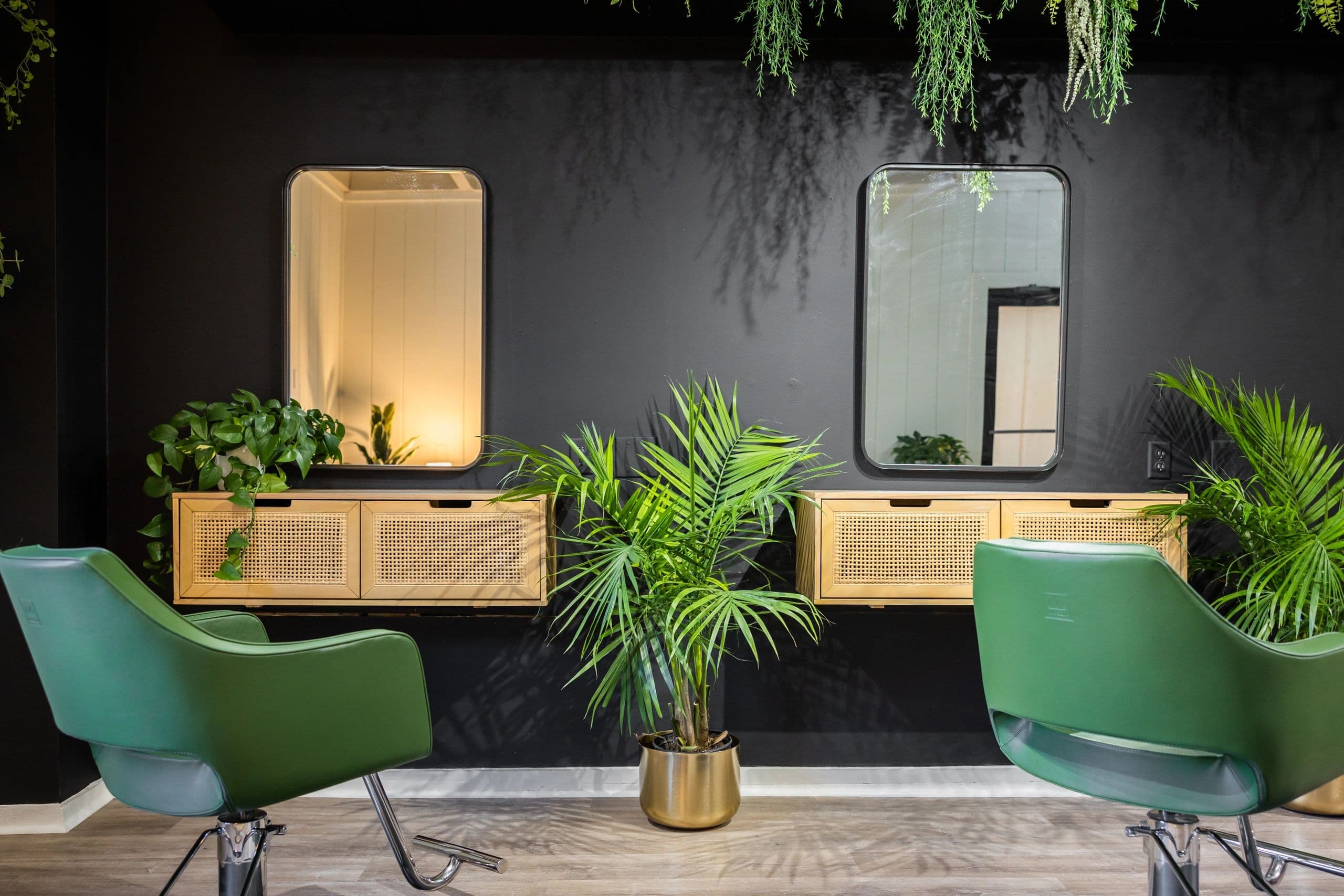 Styling stations with green chairs, rattan consoles, and hanging greenery against a dark accent wall